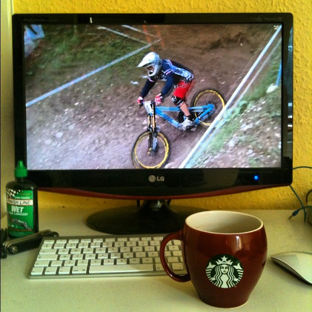 Computer monitor showing a cyclist on a dirt trail, with a keyboard, mouse, and brown Starbucks mug on the desk.