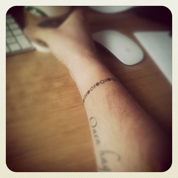 Forearm with script and dotted tattoos resting on a wooden desk beside a keyboard and white mouse.