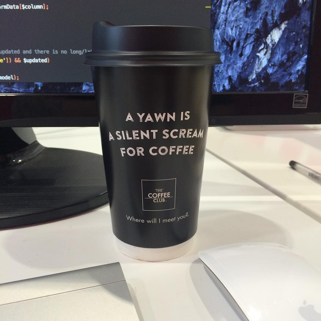 Black takeaway coffee cup on a desk in front of computer monitors, printed with the text “A yawn is a silent scream for coffee.”