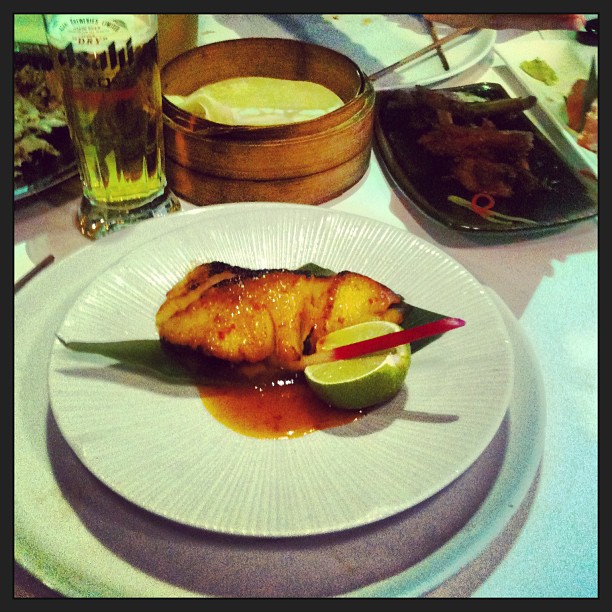 Glazed fish fillet on a white plate with lime and red garnish, beside a beer and several dishes on a restaurant table.