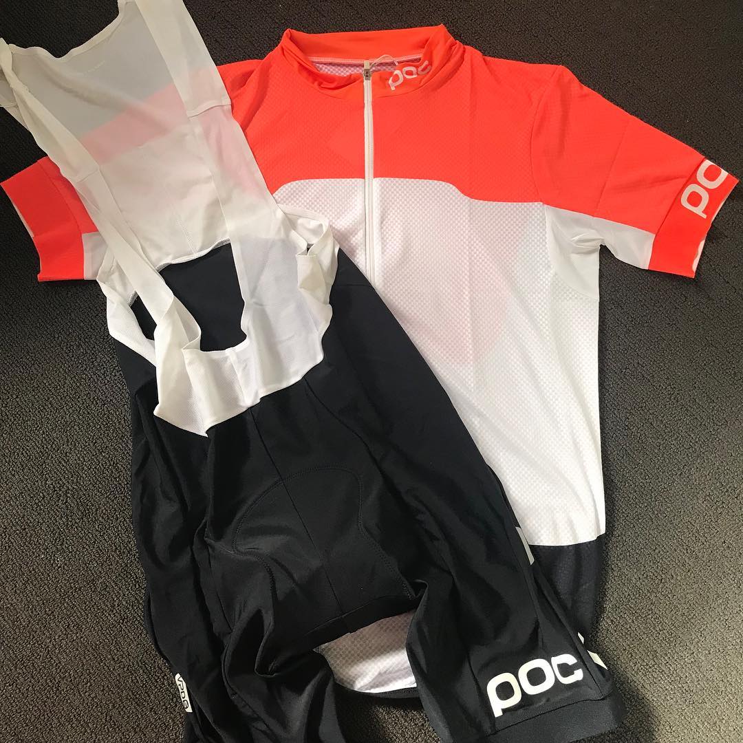 Bright coral, white, and black cycling jersey with black bib shorts laid out on a dark textured floor.