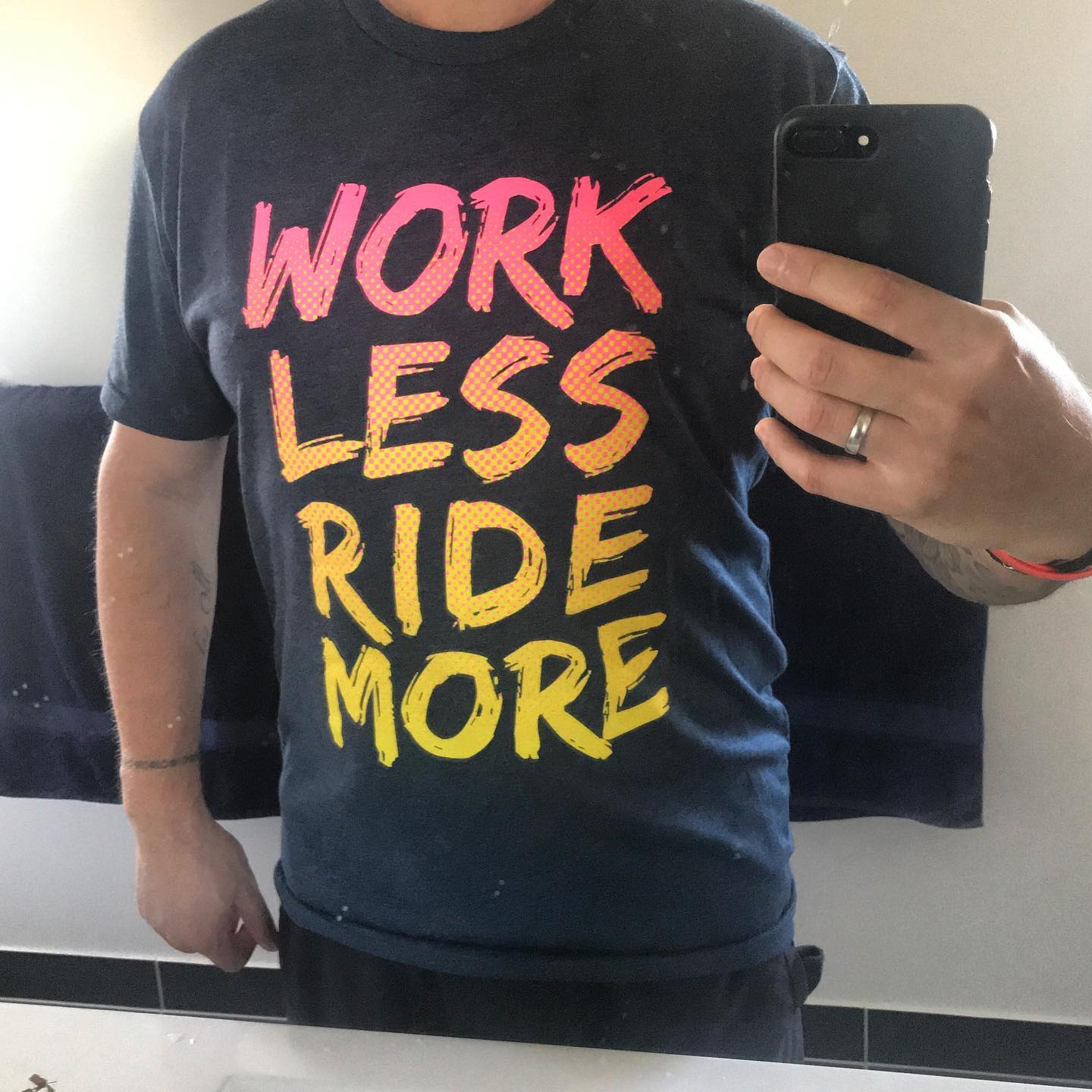 Person taking a mirror selfie in a black T-shirt with bright pink and yellow text reading “WORK LESS RIDE MORE.”