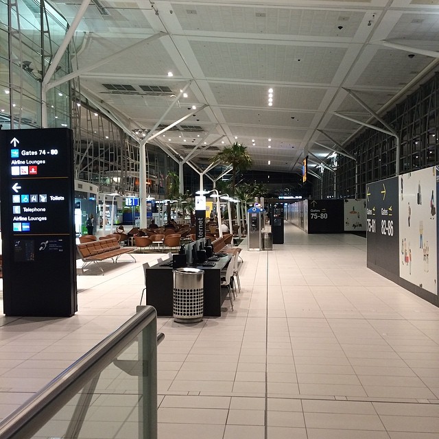 Quiet airport terminal with gate signs, seating areas, glass walls, and a wide tiled walkway under a high white ceiling.