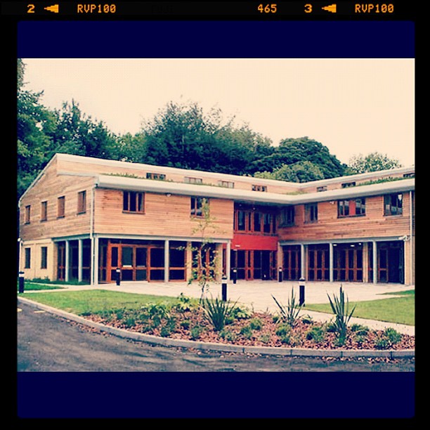 Modern two-story wood building with large windows, a red entrance, and landscaped grounds bordered by trees.
