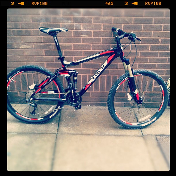 Black and red Trek mountain bike leaning against a brown brick wall on a paved sidewalk.