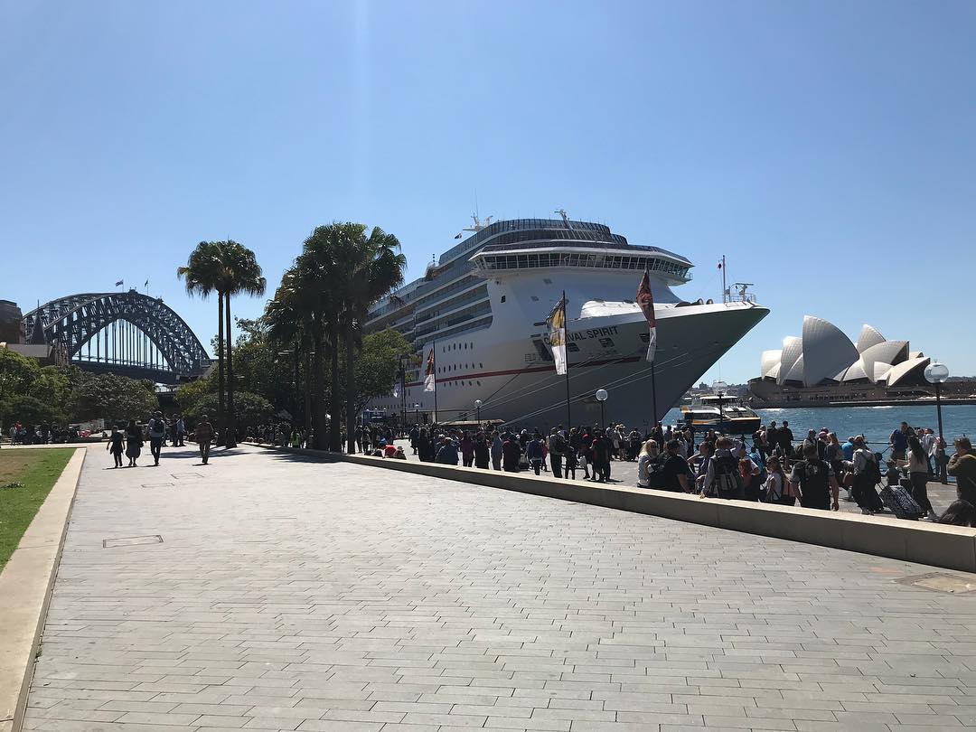Crowd along a sunny waterfront promenade beside a large cruise ship, with Sydney Harbour Bridge and the Opera House in the background.