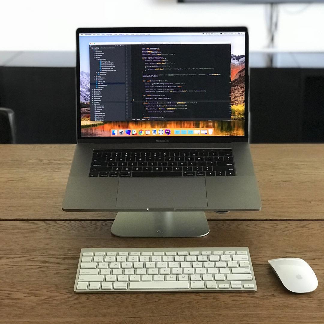 Laptop on a stand displaying code, with a wireless keyboard and mouse on a wooden desk.
