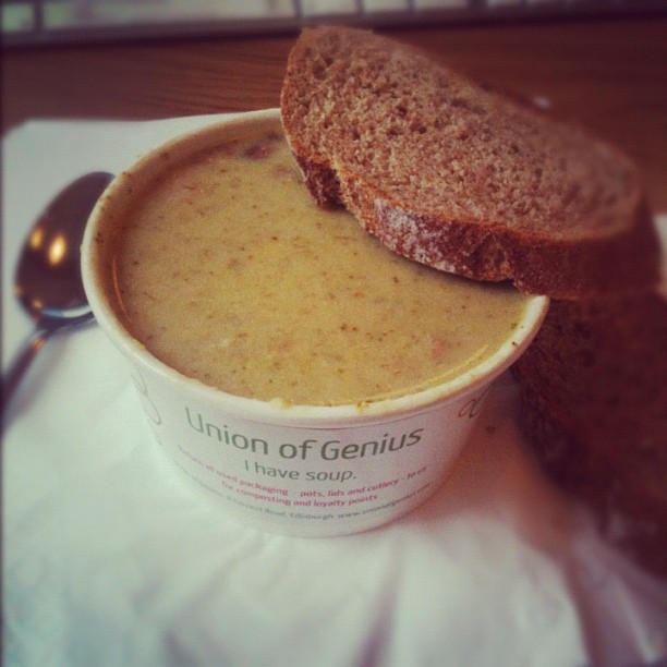 A paper cup of creamy soup with two slices of brown bread on top and a spoon beside it.