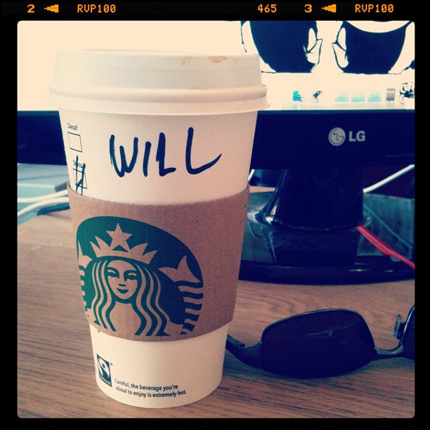 Starbucks coffee cup labeled "WILL" on a desk in front of an LG monitor, with sunglasses beside it.