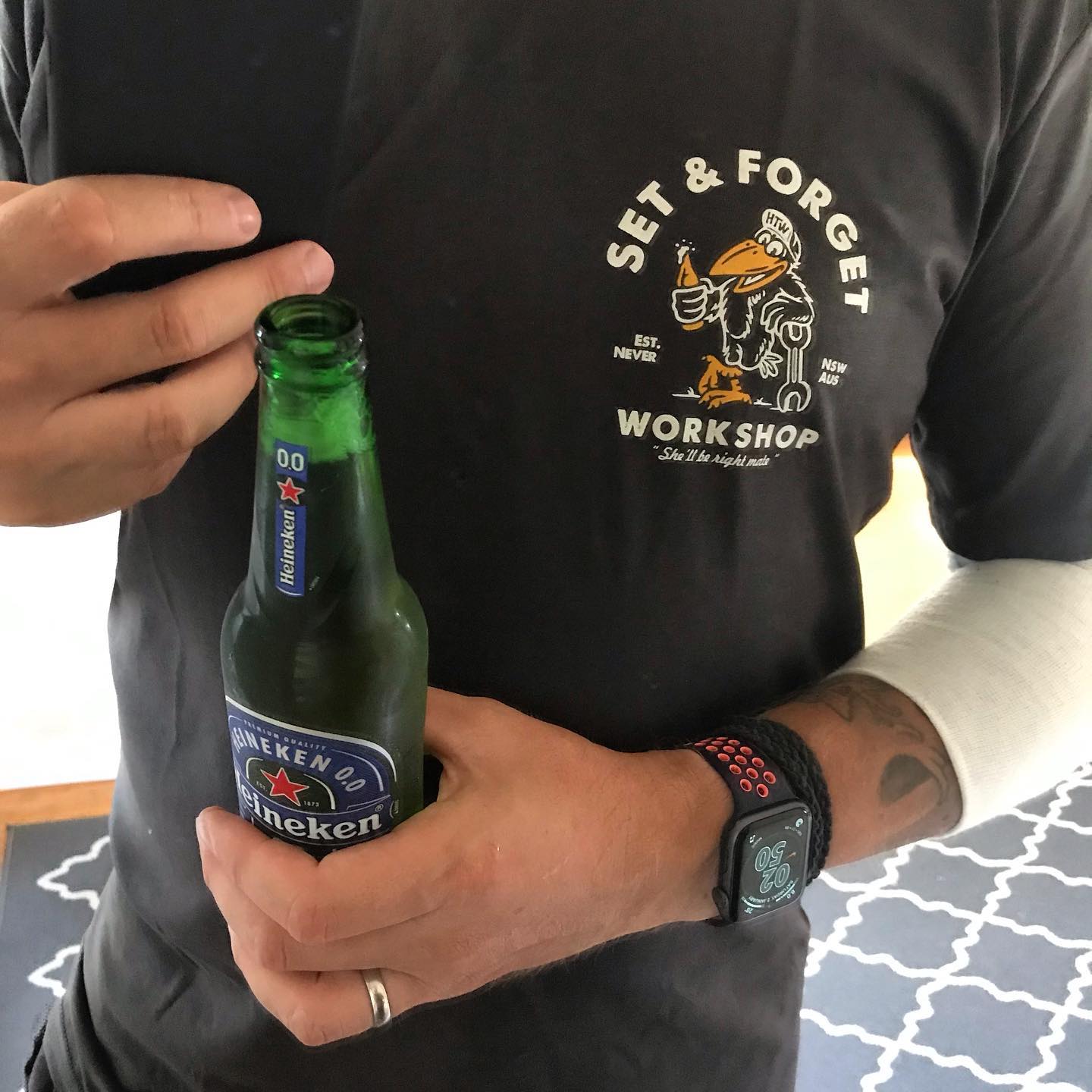 Person in a black shirt holding a Heineken 0.0 bottle, wearing a smartwatch and a white cast on one arm.