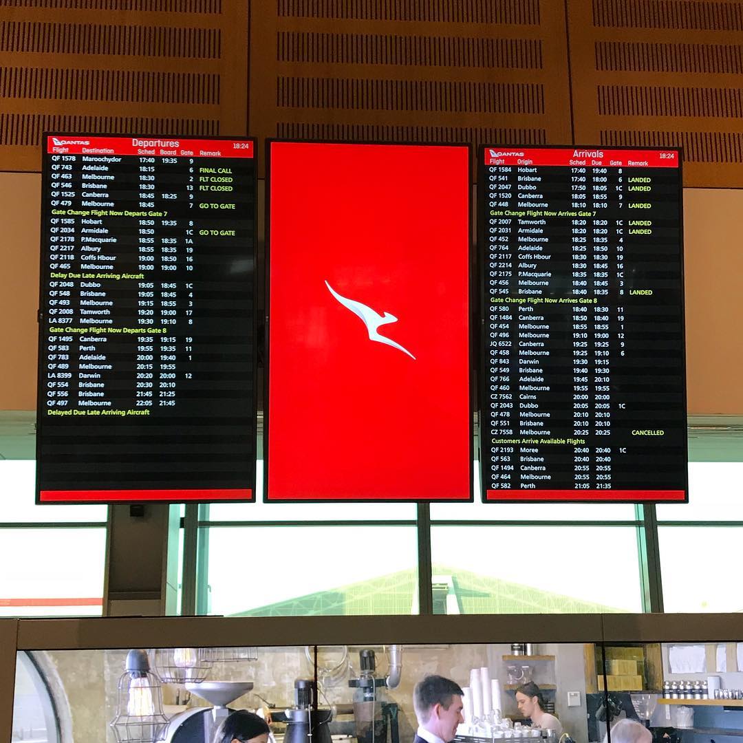 Three airport display screens show Qantas departures and arrivals, with a red kangaroo logo on the center screen above a café counter.