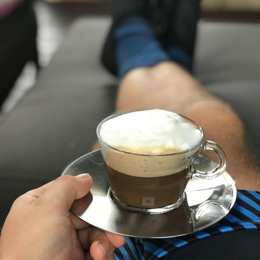 Hand holding a glass cup of foamy coffee on a metal saucer, with a person’s legs stretched out on a couch in the background.