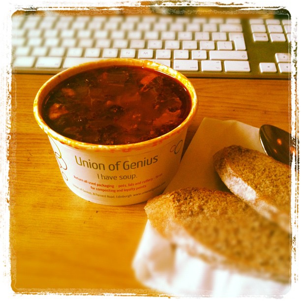 Cup of dark red soup on a desk beside bread rolls and a spoon, with a computer keyboard in the background.