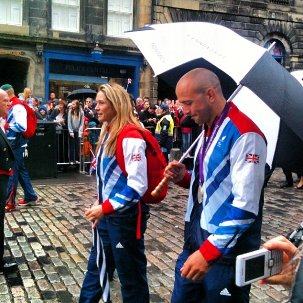 Two athletes in Team GB tracksuits walk on a wet cobblestone street, one holding a large black-and-white umbrella and a medal.