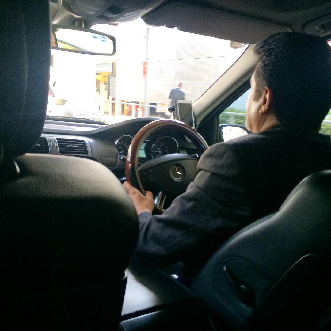 View from the back seat of a car showing a driver at the wheel, dashboard, and a bright street scene through the windshield.