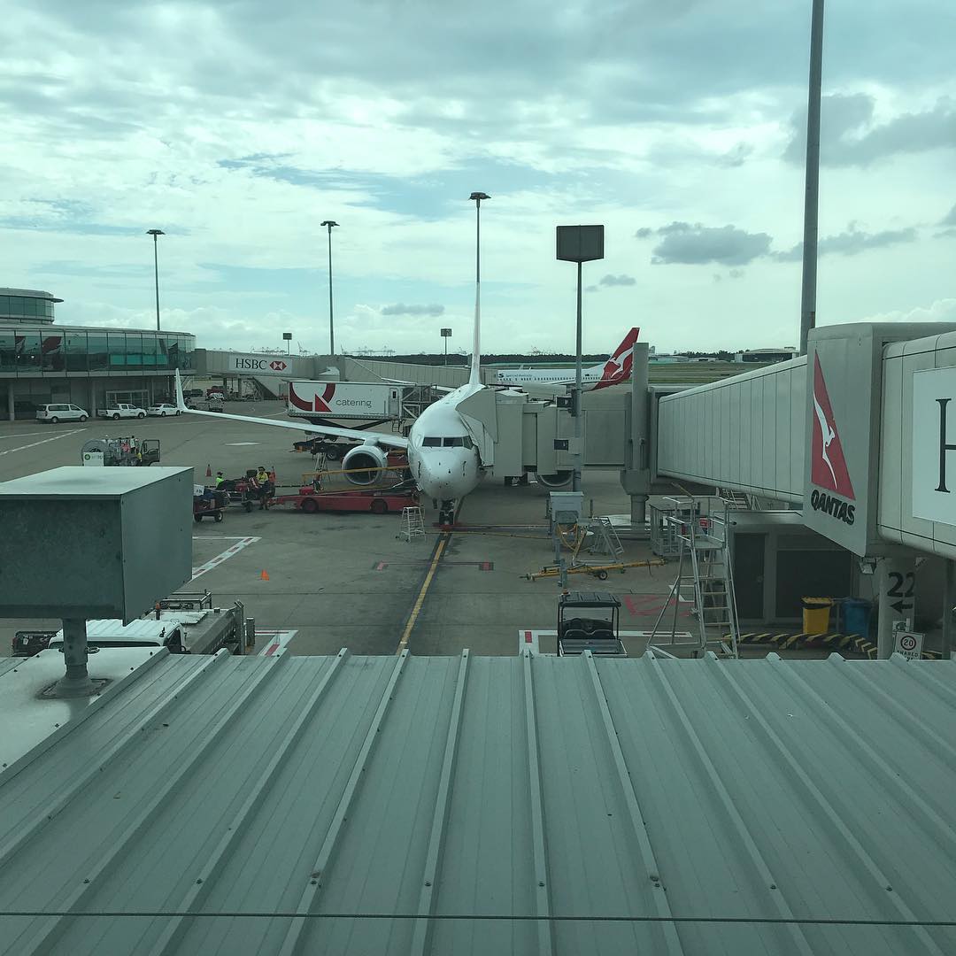 Qantas airplane parked at an airport gate with a jet bridge attached, ground vehicles nearby, and a cloudy sky overhead.