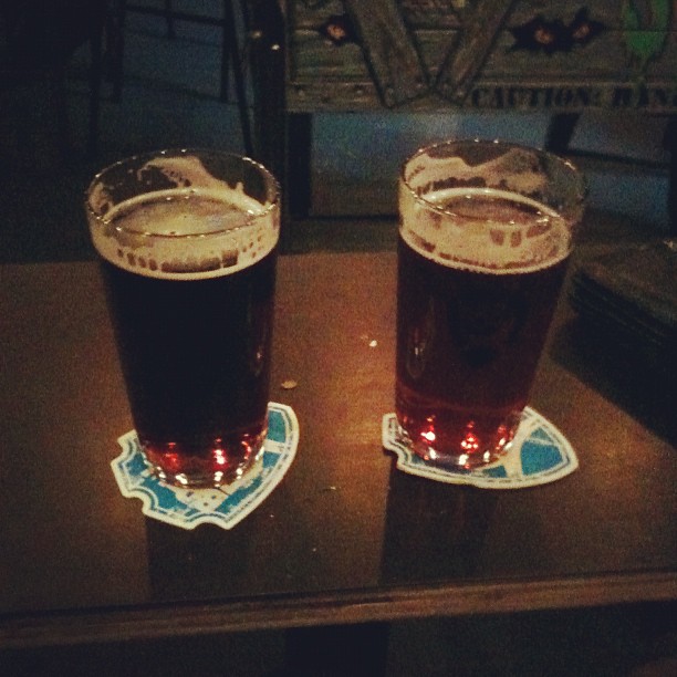 Two glasses of dark beer on blue coasters on a dimly lit table.