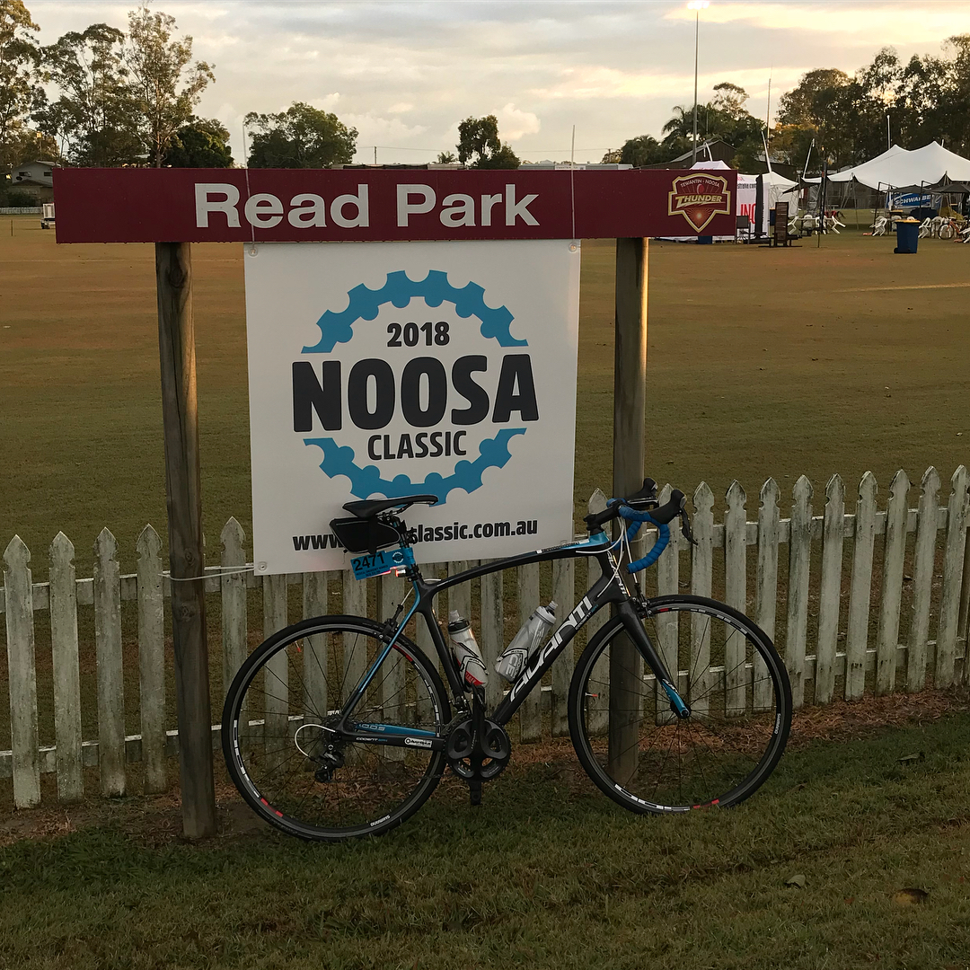 Road bike leaning against a white picket fence beneath a Read Park sign and a 2018 Noosa Classic banner on a grassy field.