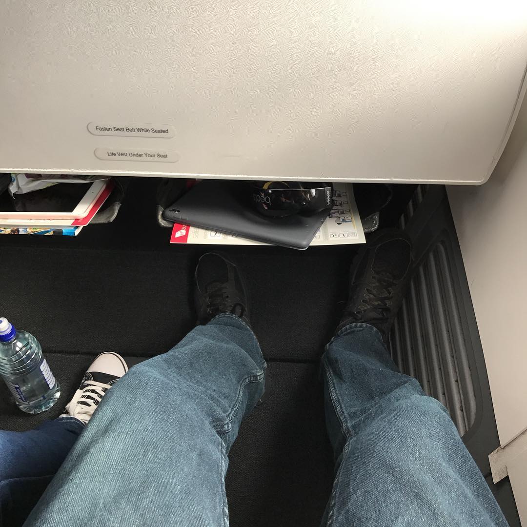 View from an airplane seat showing extended legroom, blue jeans, black shoes, a seat pocket with a tablet and headphones, and a water bottle.