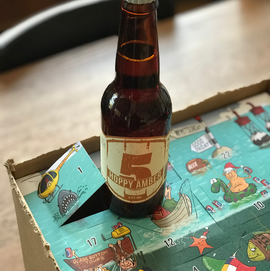 Brown beer bottle labeled "5 Hoppy Amber" standing in a cardboard advent-style box with colorful illustrated numbered flaps.