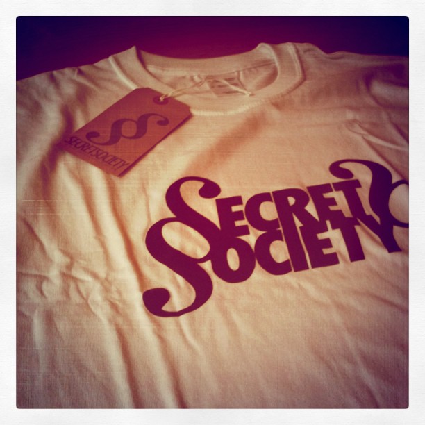 White T-shirt with purple “Secret Society” text and a hanging tag on the collar.