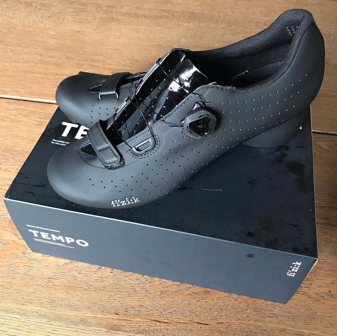 Pair of black cycling shoes with perforated uppers and strap closures resting on a black Tempo shoebox on a wooden floor.