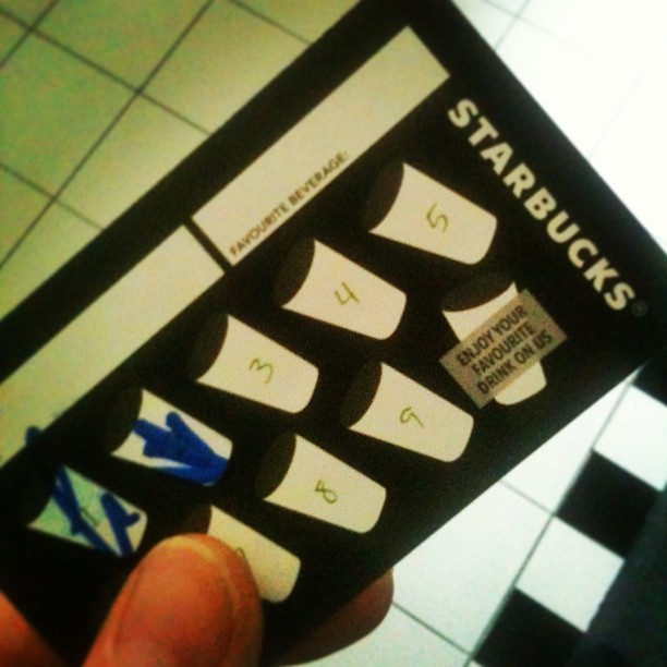 Hand holding a Starbucks loyalty card with several cup icons marked by blue Xs over a tiled floor.