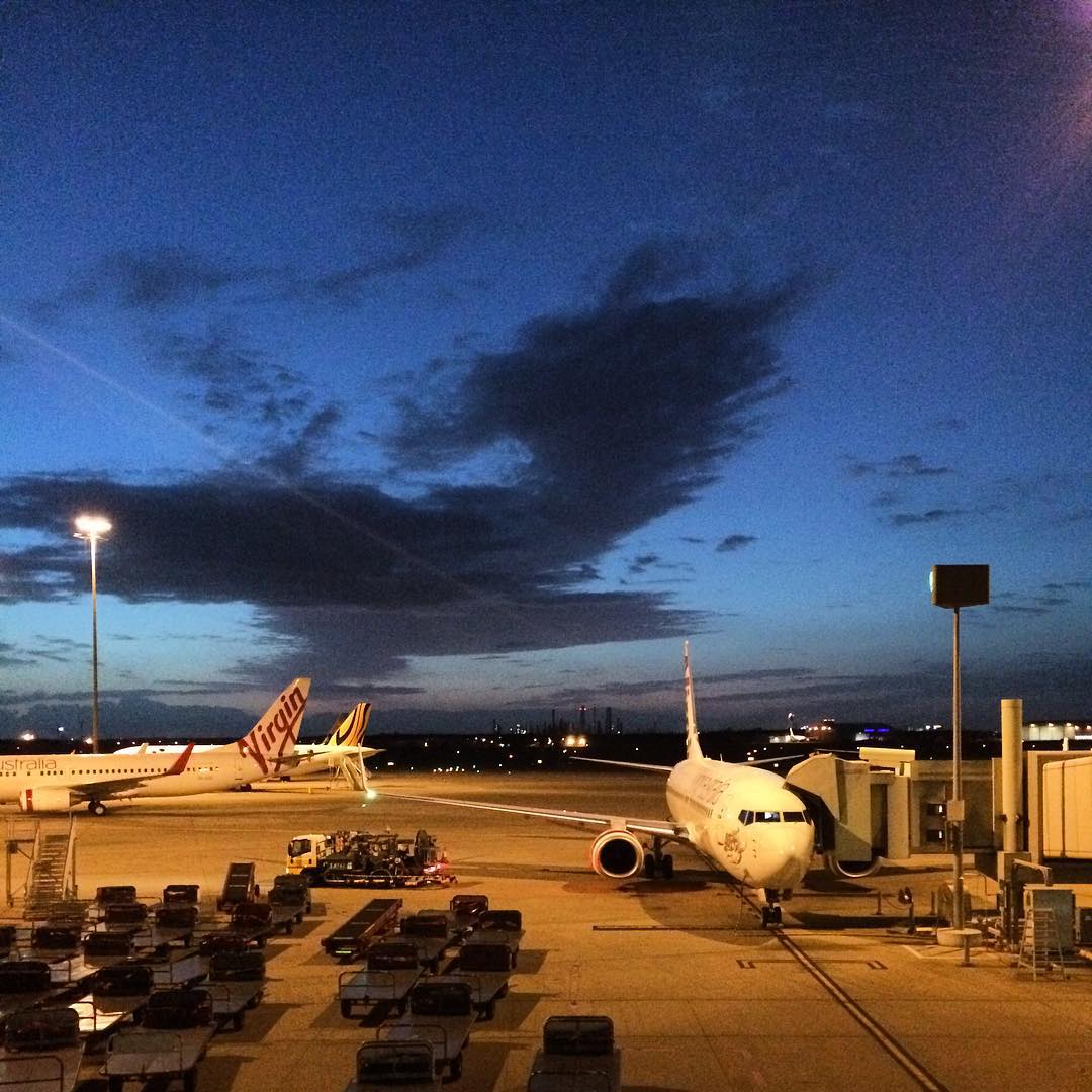 Two airplanes parked at airport gates at dusk, with baggage carts on the tarmac and dark clouds across a blue sky.