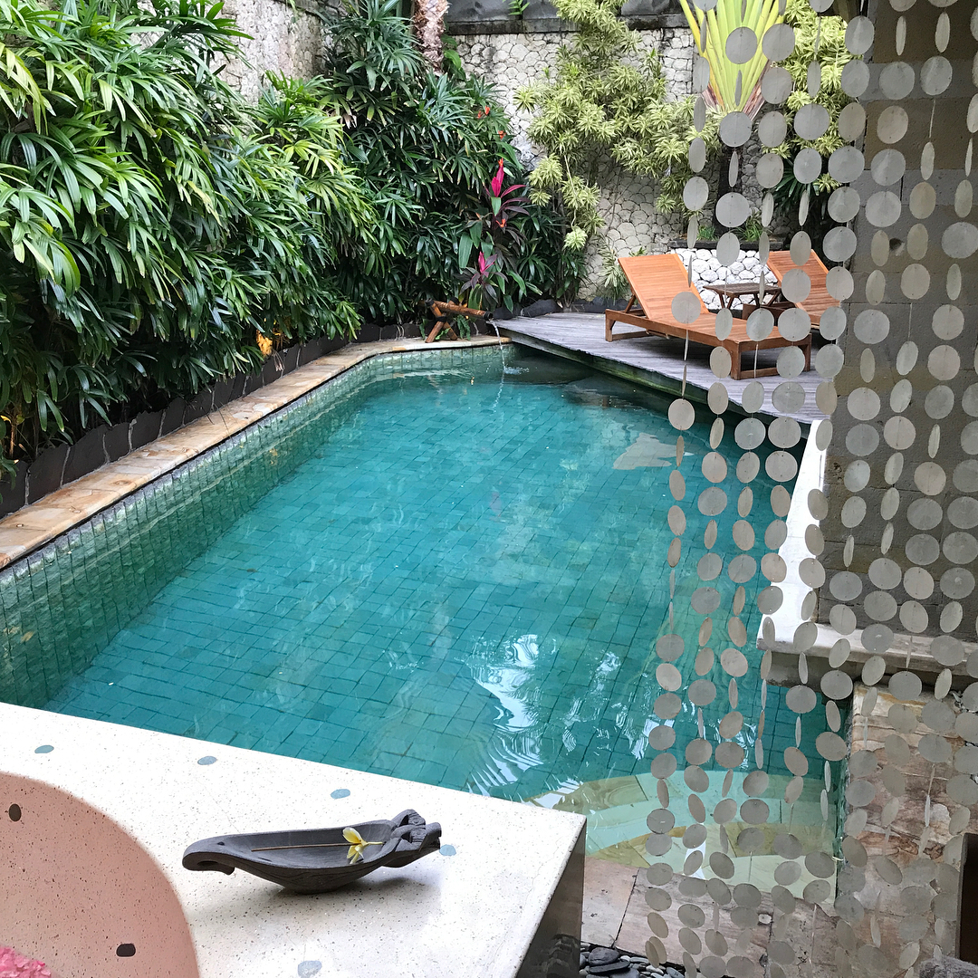Small turquoise pool beside lush tropical plants, with two wooden lounge chairs on a deck and a hanging bead curtain in the foreground.