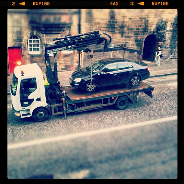 Tow truck with a crane lifting a black sedan onto its flatbed on a cobblestone street beside a stone building.