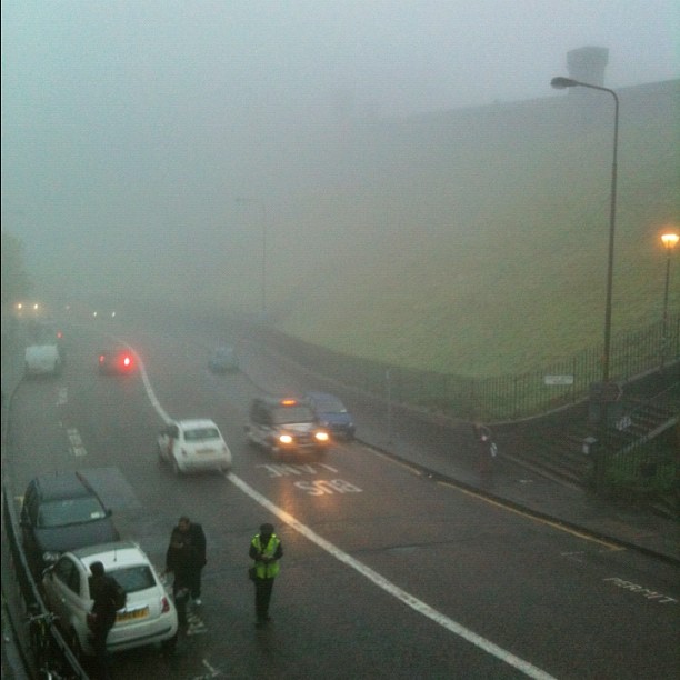Foggy street with several cars, two pedestrians, and a grassy hillside beside a sidewalk and streetlights.