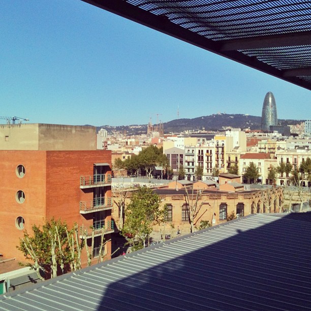 City skyline with low-rise buildings, a tree-lined street, distant hills, and a bullet-shaped tower under a clear blue sky.