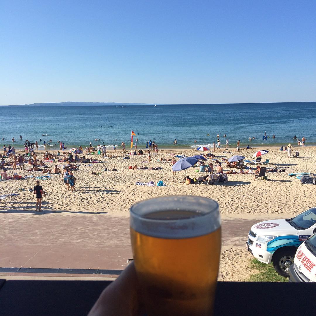 Glass of beer in the foreground overlooking a crowded sandy beach, calm blue ocean, umbrellas, swimmers, and a clear sky.