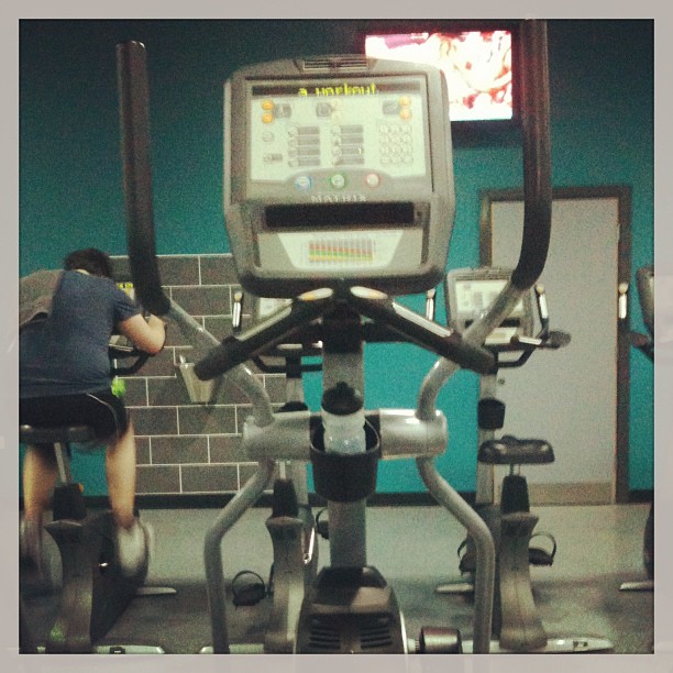 Front view of an elliptical machine in a gym, with a person exercising on a bike in the background.