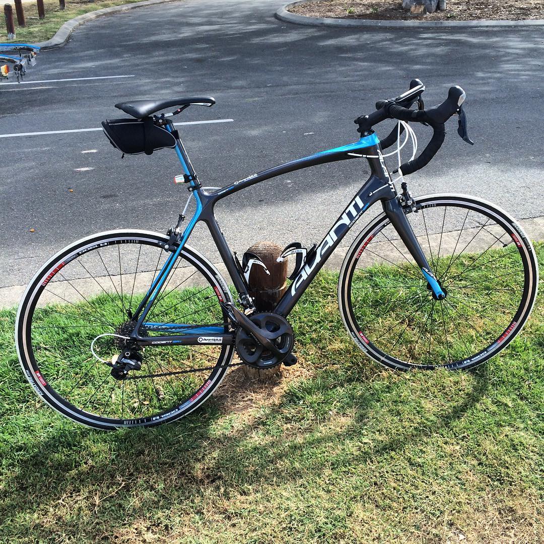 Black and blue road bike with drop handlebars and a small saddle bag, leaned against a short wooden post beside a paved lot.