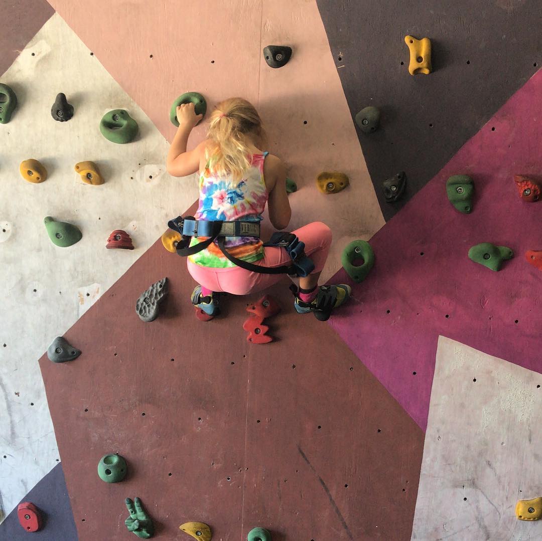 Child in a tie-dye tank top and pink leggings climbs an indoor rock wall while wearing a harness.