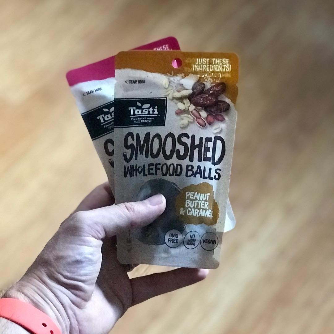 Hand holding two Tasti Smooshed Wholefood Balls snack pouches, including peanut butter and caramel, against a wooden floor.