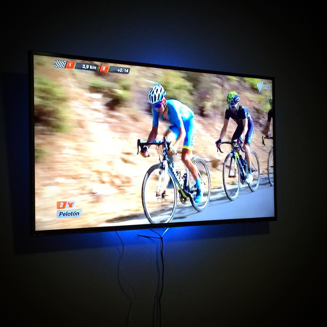 Wall-mounted TV showing a cycling race with two riders on a road, blue backlight glowing behind the screen and cords hanging below.
