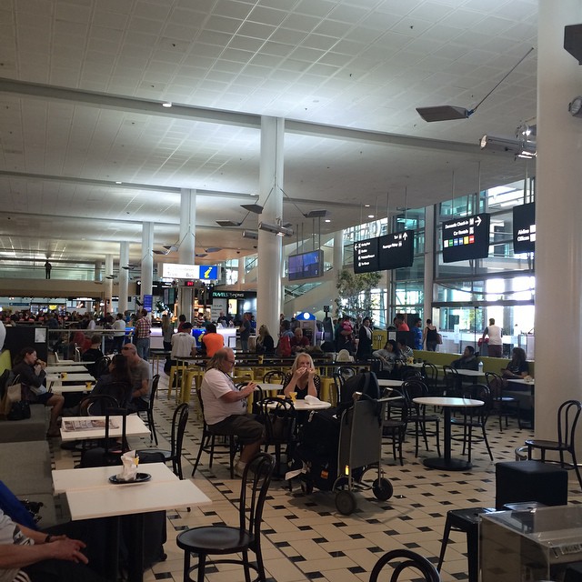 Airport terminal seating area with café tables and chairs, travelers sitting and walking, and overhead departure signs near large windows.