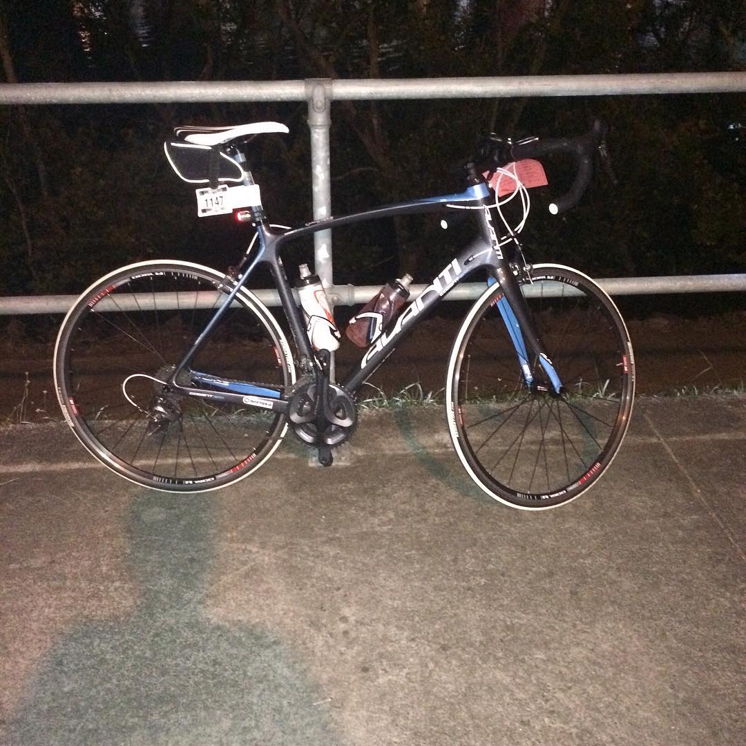 Black and blue road bike with drop handlebars, two water bottles, and a race number tag parked by a metal railing at night.