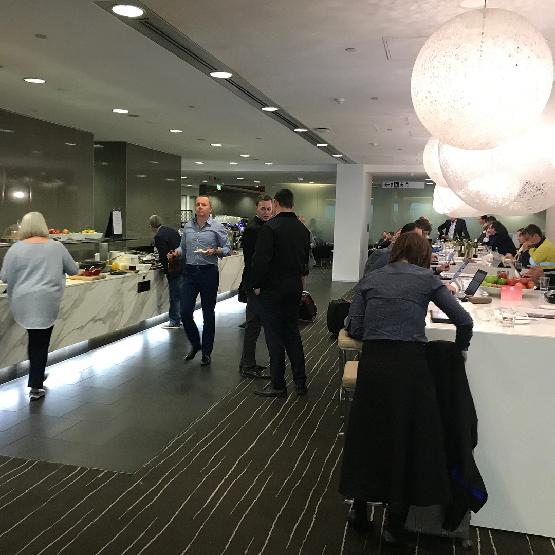 Modern airport lounge with people eating, walking, and working at a long counter under large glowing pendant lights.
