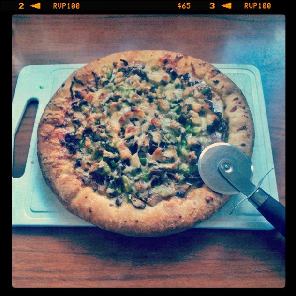 Round pizza on a white cutting board with a pizza cutter, topped with melted cheese, green peppers, and mushrooms.