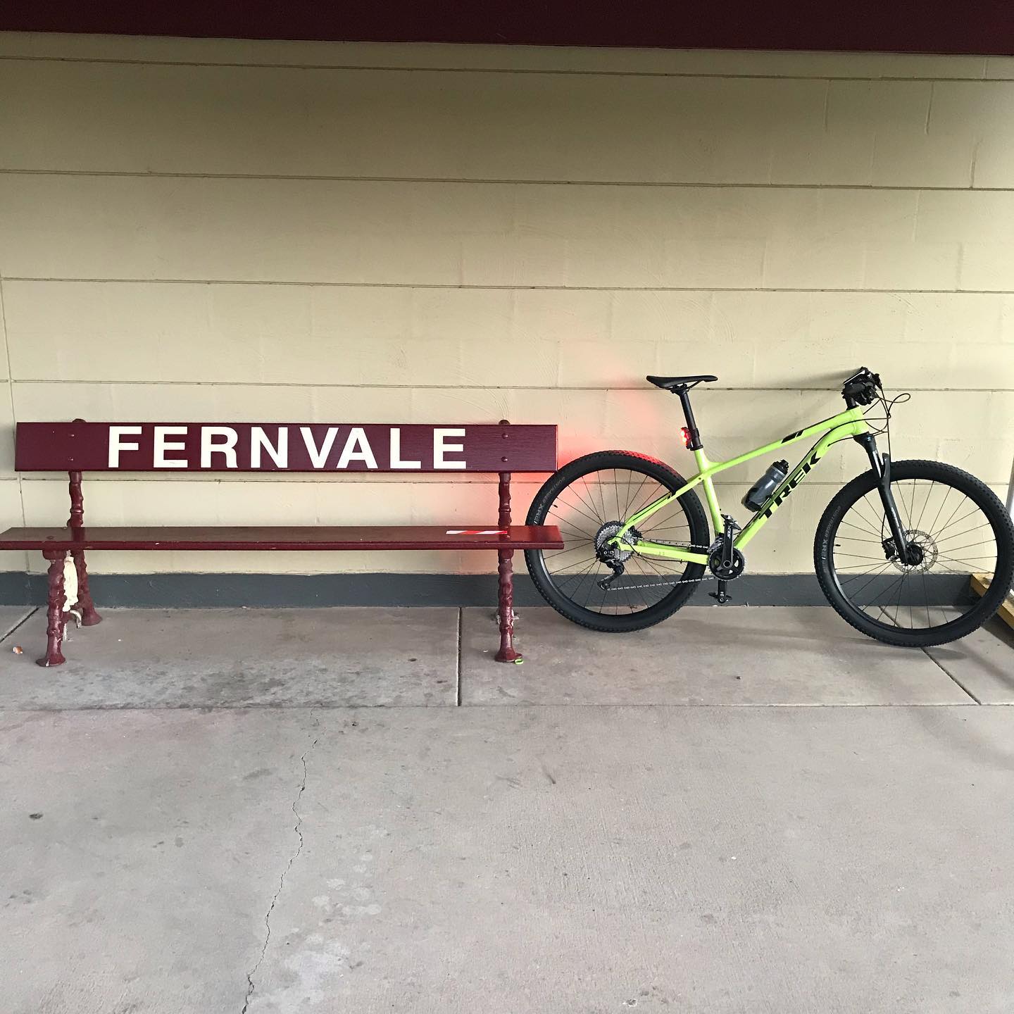 Maroon bench with “FERNVALE” painted in white beside a bright green bicycle against a cream brick wall.