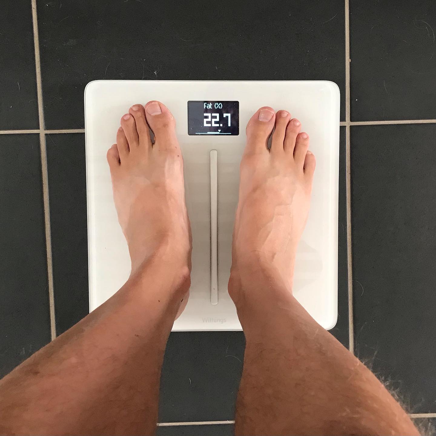 Bare feet on a white digital bathroom scale on dark tile, with the display showing body fat at 22.7%.