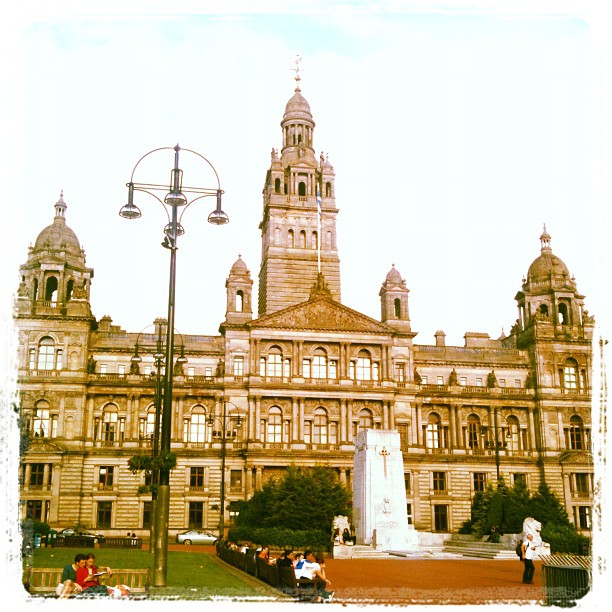 Historic stone building with a central clock tower and domed corners, seen across a plaza with a monument, lamppost, and people seated nearby.