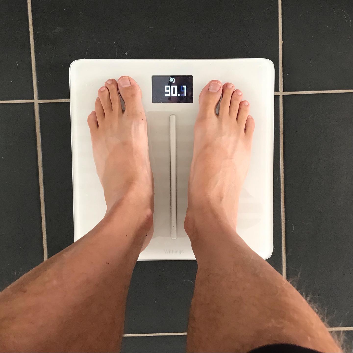 Bare feet standing on a white digital bathroom scale displaying 90.9 kg on a dark tiled floor.