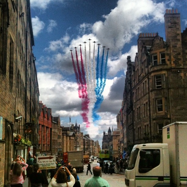 Jets flying in formation trail red, white, and blue smoke above a city street lined with tall stone buildings and onlookers.