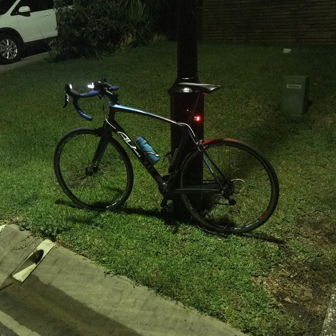 Road bike with blue handlebar tape leaning against a black pole on grass at night.