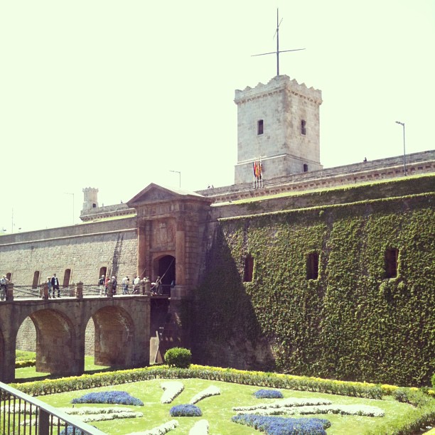 Stone fortress with an ivy-covered wall, arched bridge, clock-shaped garden, and a square tower with a flag above the entrance.