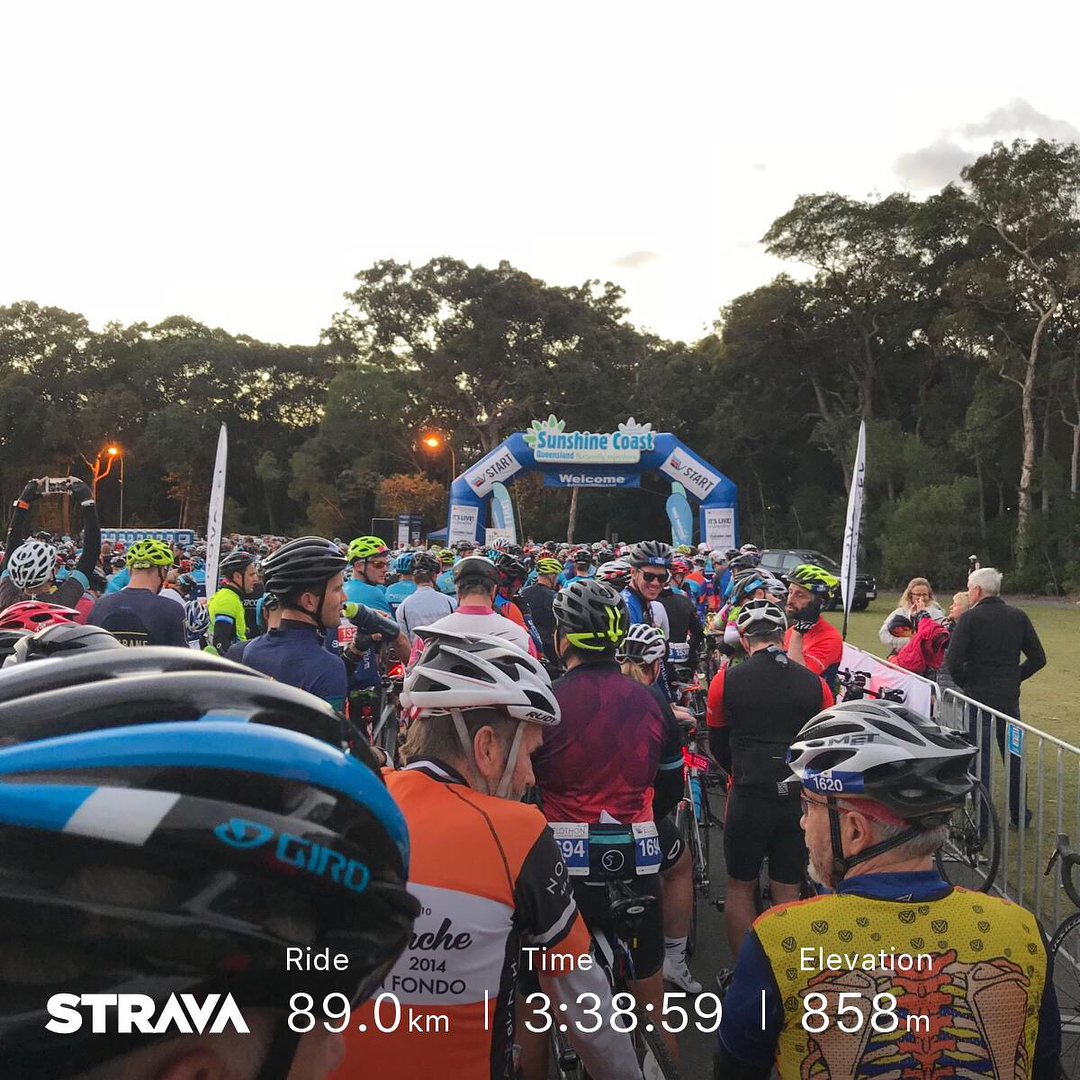 Crowd of cyclists in helmets gathered at a Sunshine Coast start arch, with Strava stats overlaid at the bottom.